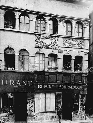 La même façade en 1921, photographiée par Henri Heuzé pour les Monuments historiques. Le décor de pierre est intact. Sur la devanture, peint en grandes lettres claires : « Cuisine bourgeoise », et sur la vitrine « Restaurant ». Un enfant se tient debout sur le trottoir, à droite de la porte.