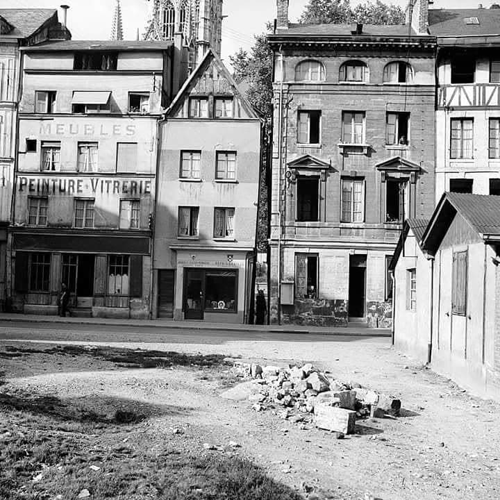 Photographie d'après-guerre de la rue Eau-de-Robec. Au premier plan, un terrain vague en terre battue avec un tas de moellons et de pierres de taille. De l'autre côté de la rue, des maisons debout mais abîmées, dont une portant les inscriptions peintes « Meubles » et « Peinture-Vitrerie », et une baraque de bois à droite. Au fond, les flèches de la cathédrale de Rouen.