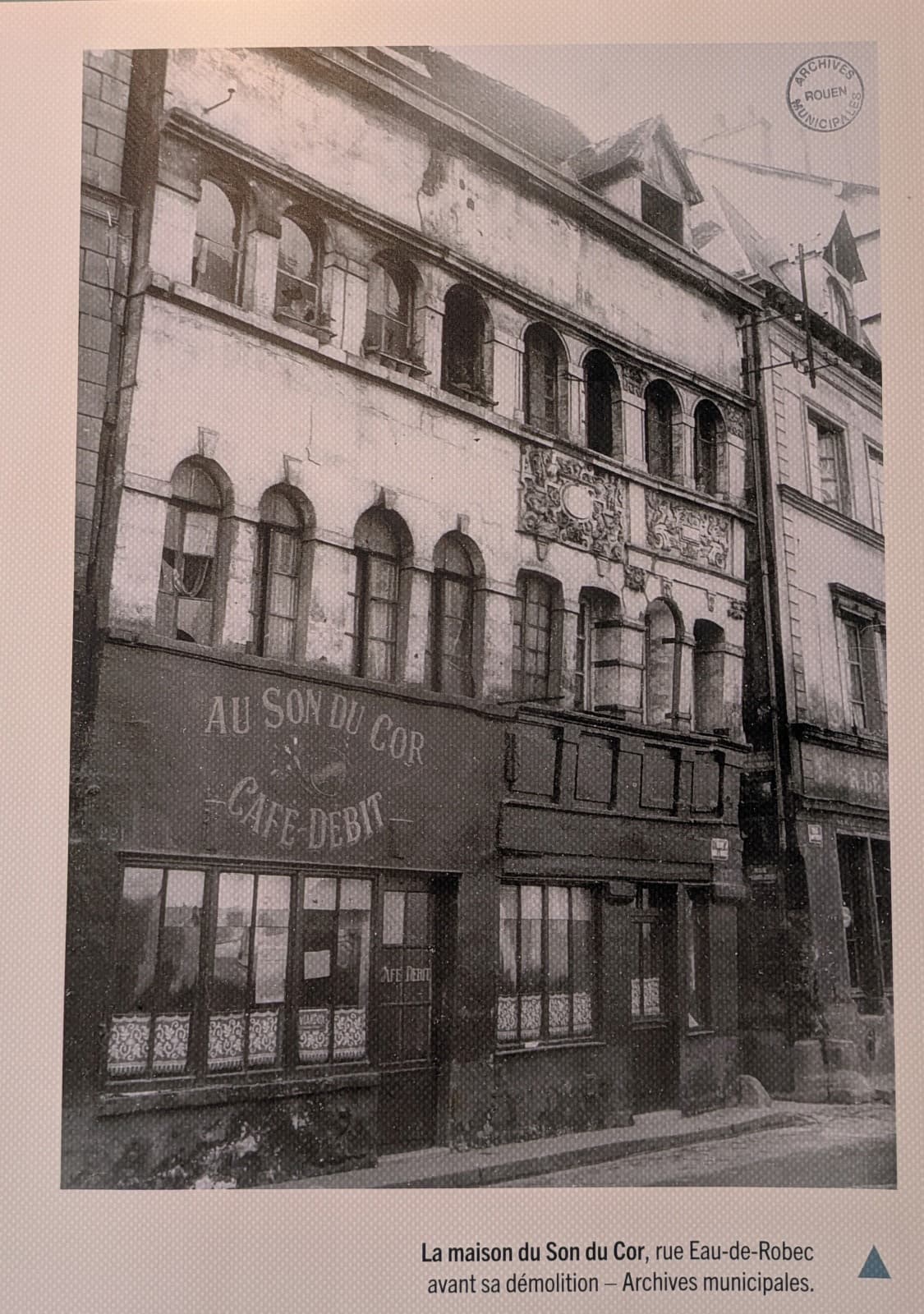 La maison vue en oblique depuis la rue, avant sa démolition. Sur le bandeau sombre de la devanture, peint en lettres claires : AU SON DU COR, un cor de chasse dessiné en dessous, puis CAFÉ-DEBIT. Au-dessus, les baies en plein cintre et les deux frises sculptées de la maison Renaissance.
